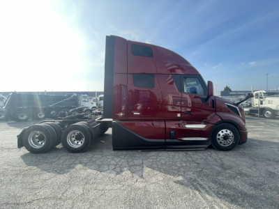 Side view of a maroon Volvo VNL860 conventional sleeper truck tractor with a full sleeper cab in a dealership lot. Visible text on the cab includes no legible slogans; appears to be new with triple rear axles and chrome exhaust, low front b
