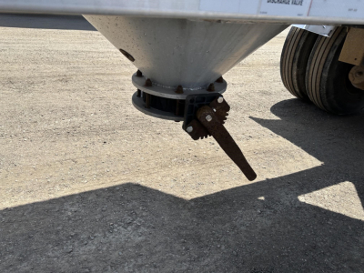 Pneumatic trailer underside showing conical hopper and rusted discharge chute, wheel set in background, gravel surface.