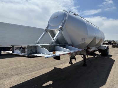Trailer viewed from rear left side showing cylindrical tanker with arched landing gear and support frame; small label on tank; dry bulk pneumatic trailer in a yard.