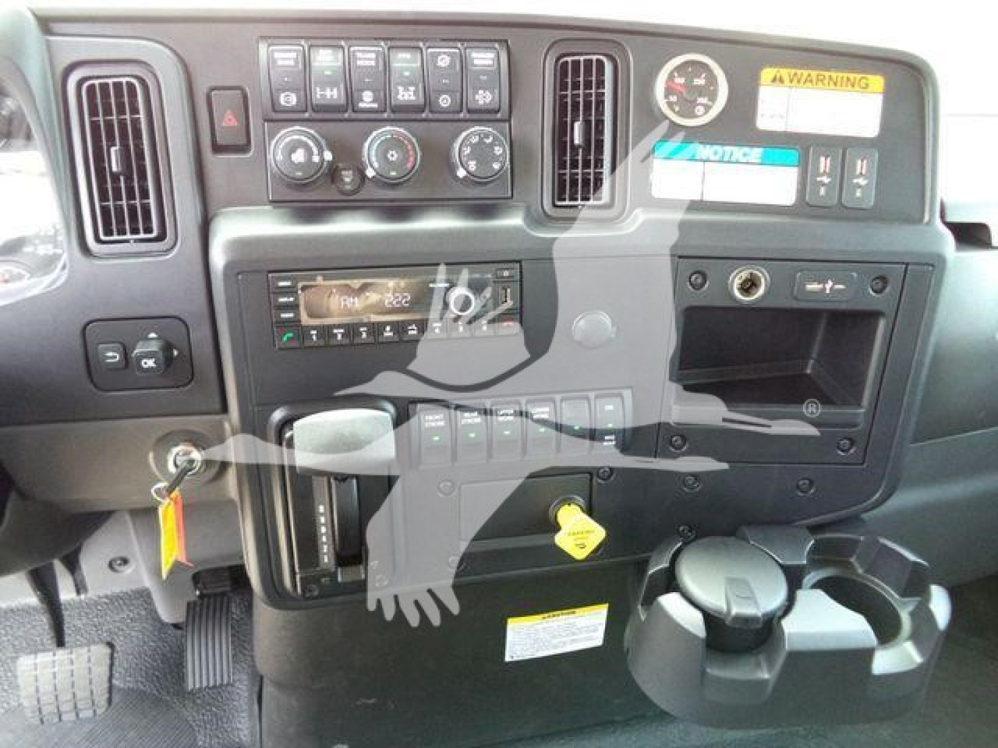 Dashboard and controls of an International MV truck cab showing ignition, steering area, climate controls, cup holders, and a yellow key, with warning stickers visible.