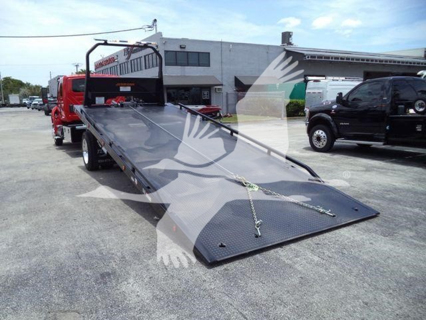 Truck bed with a lowered flatbed tow truck in a dealership lot showing a black diamond-plate ramp and chain ties.