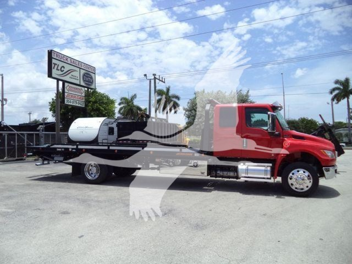 Truck in a lot with a red cab and a flatbed rollback bed, monochrome sky, dealership sign in background, VIN and stock number not visible on text.