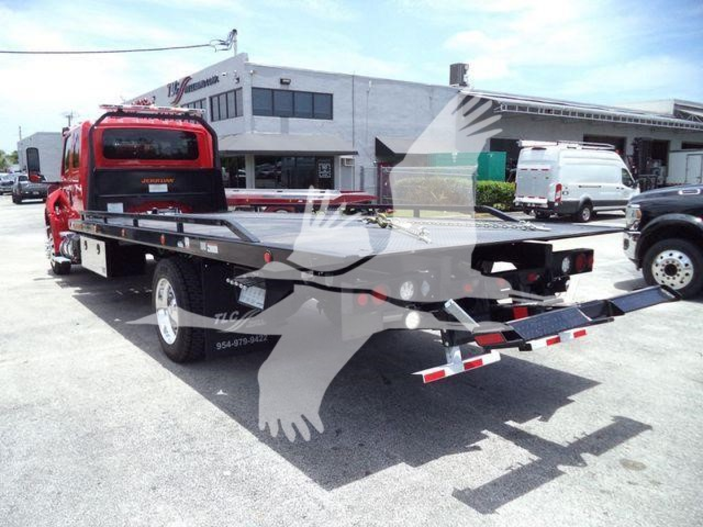 Truck bed of a rollback tow truck with a red cab and black flatbed, viewed from rear-left; VIN and stock number not visible; service yard with buildings in background.