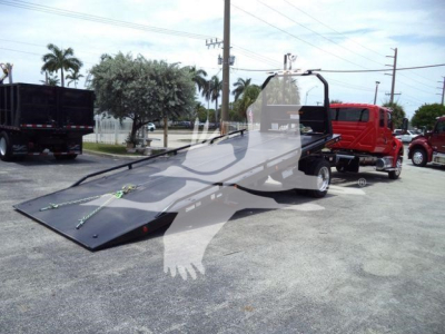Truck rollback tow truck with extended flatbed in driveway. Red tow truck in background, trees and power lines, asphalt surface.