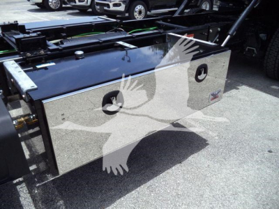 Aluminum toolbox mounted on a truck chassis with two circular latches and a logo plate; reflective surface shows ground and other equipment in background.