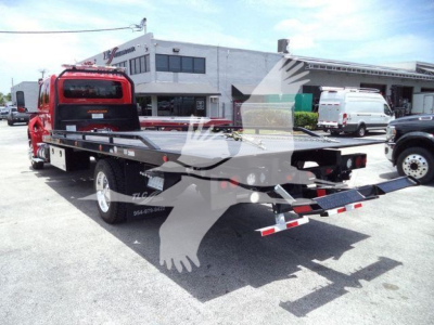 Truck bed of a rollback tow truck with a red cab and black flatbed, viewed from rear-left; VIN and stock number not visible; service yard with buildings in background.