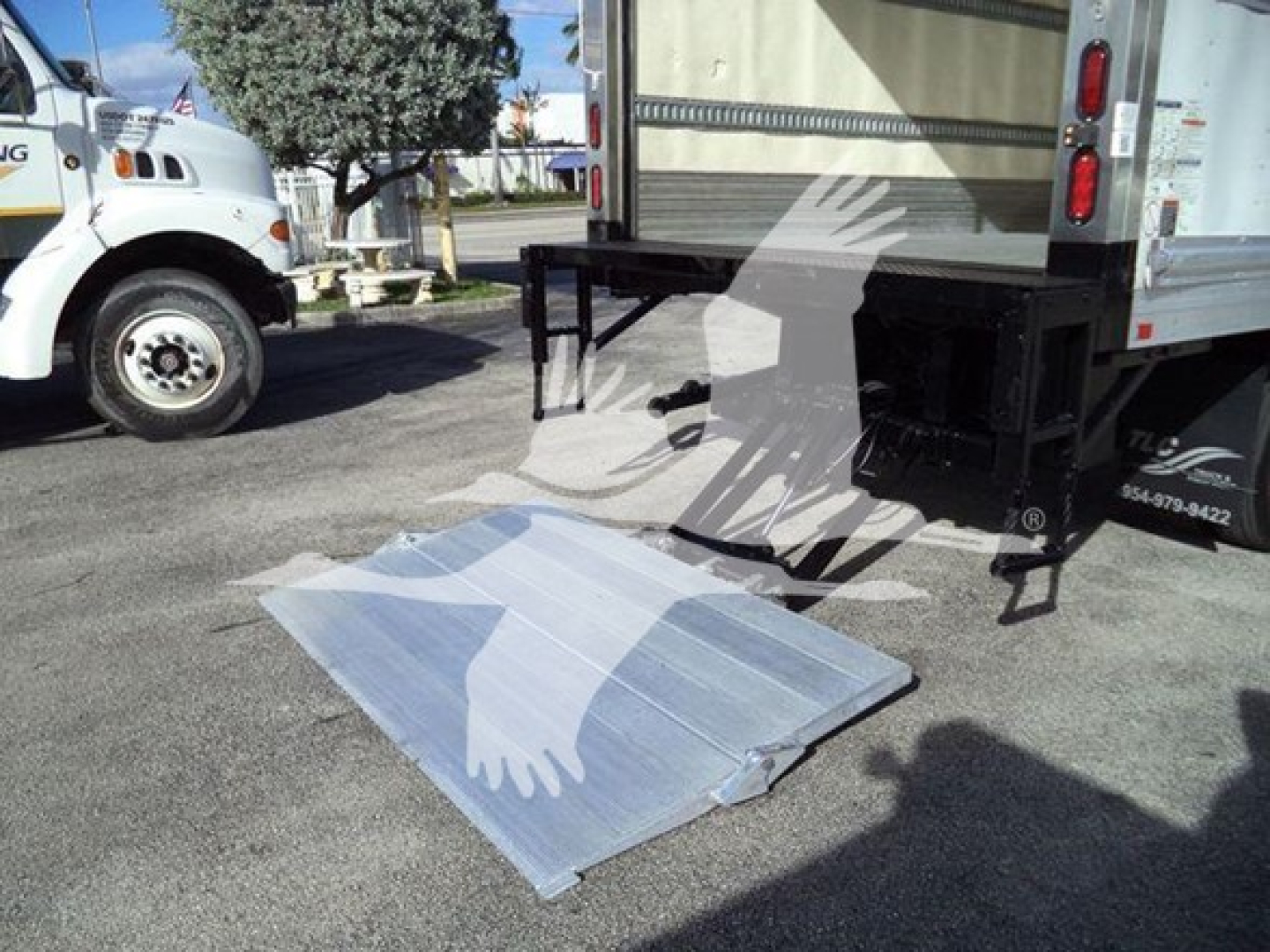 Back of refrigerated truck with unfolded aluminum ramp on ground and white pickup in background.