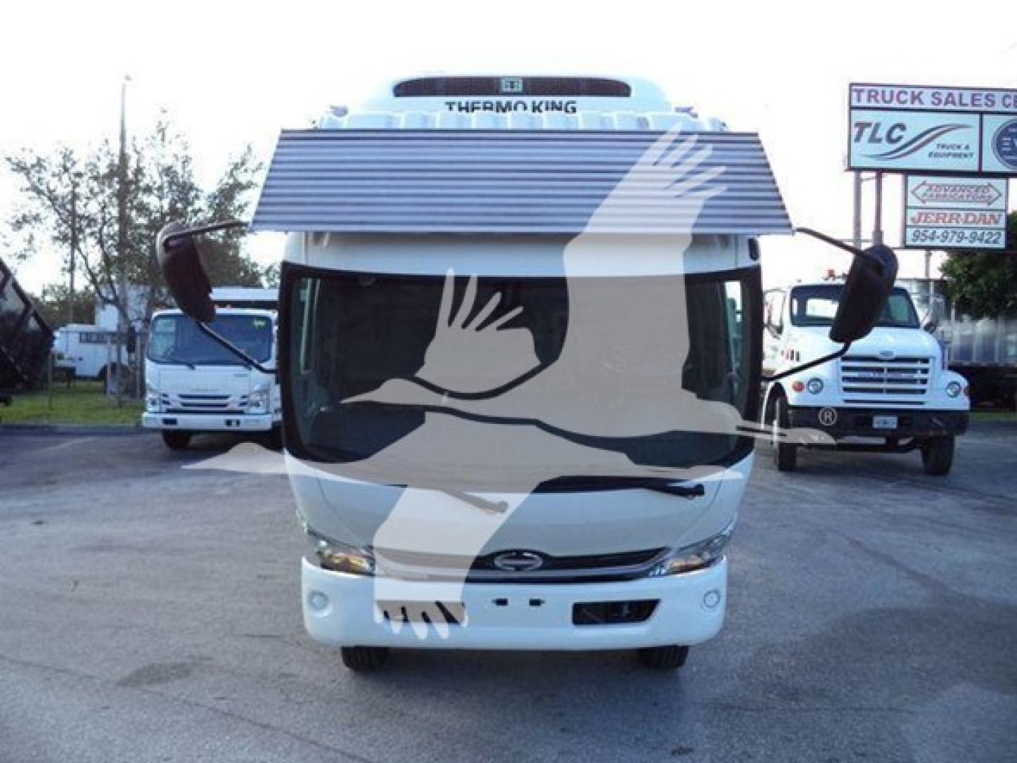 Front view of a white 2020 Hino 195 refrigerated truck with large windshield, grille, and side mirrors; dealership signs and other trucks in background.
