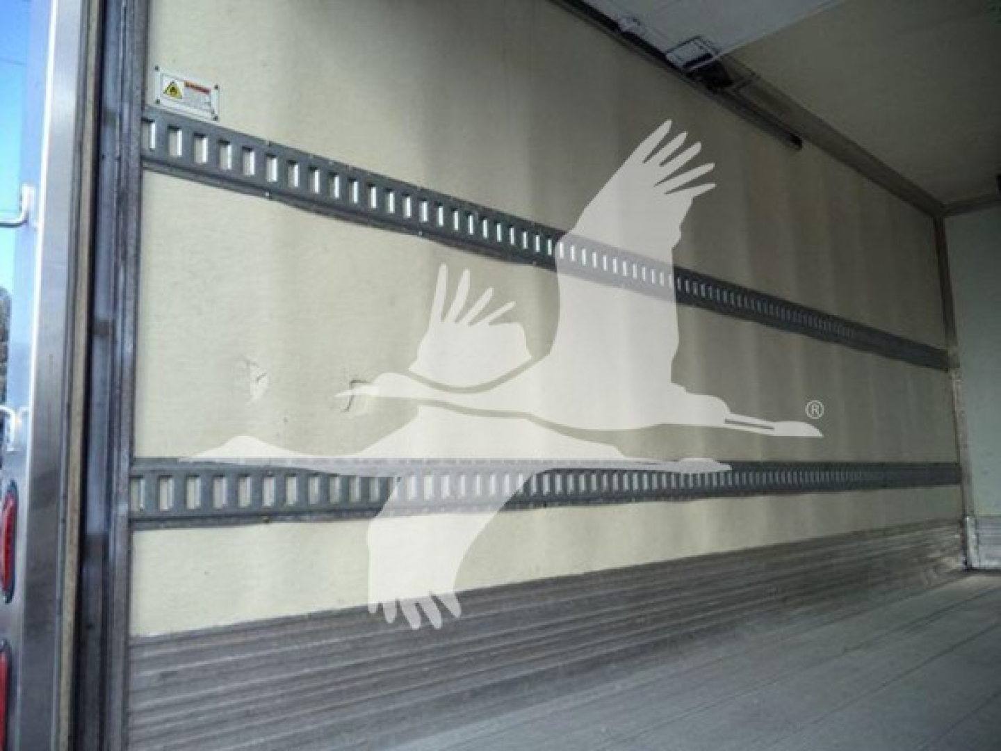 Interior of a refrigerated truck cargo area with metal rails and a light-colored wall, showing an open side door and a large graphic of flying birds.