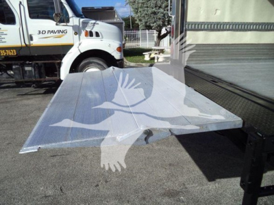 Truck with a flatbed ramp in foreground and a white Hino refrigerated truck in background. Visible text on side of truck includes 3-D PAVING and phone numbers, and the ramp edge shows corrugated metal.