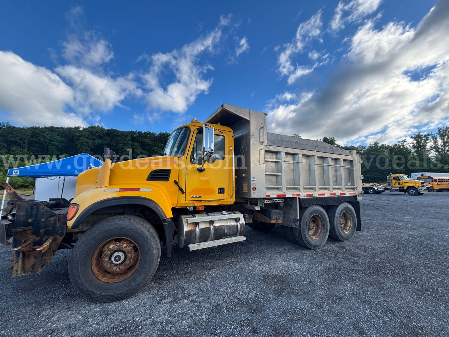 Yellow Mack CV713 dump truck with front-mounted snow plow in a gravel lot; cargo bed with gray side panels; background includes other trucks and blue sky with clouds.