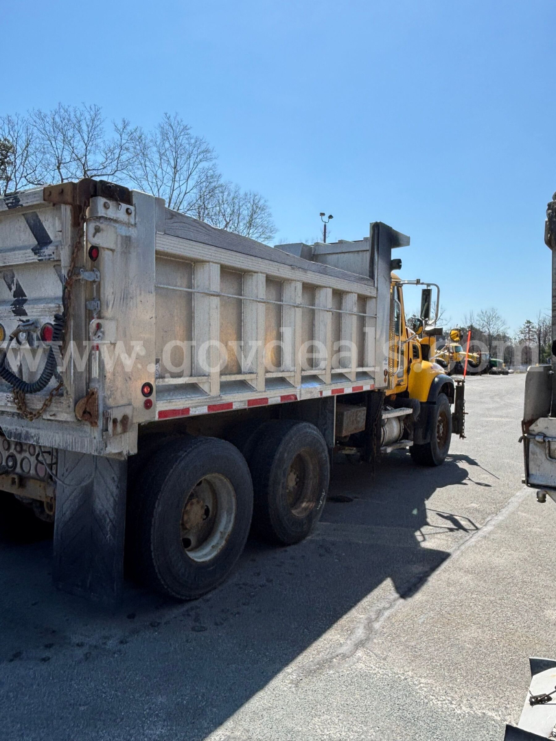 Dump truck rear left side showing cargo bed and dual rear axles, yellow tractor unit attached, with weathered metal sides and tailgate.
