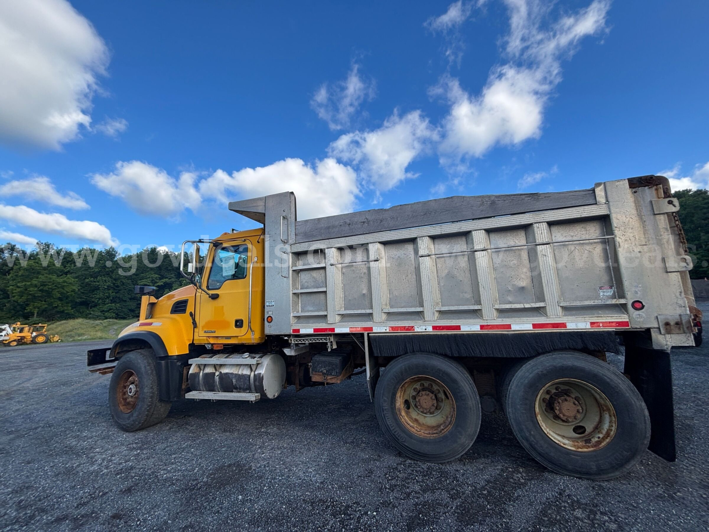 Side view of a yellow Mack CV713 dump truck with a large gray dump bed, two rear wheels, and a fuel tank; clear blue sky with clouds.