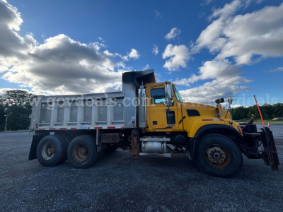 Dump truck side view of a 2004 Mack CV713 with yellow cab and silver dump bed, showing dual rear wheels and tall exhaust, under blue sky with clouds.