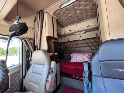 Truck sleeper cab interior showing bed with red bedspread, quilted upper panels, beige seating, and storage compartments.
