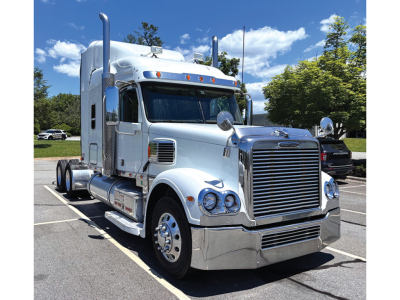 Freightliner Coronado sleeper tractor in silver/gray, front three-quarter view, chrome grille and bumper, dual side exhaust, day cab with tall sleeper area.