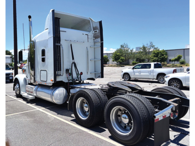 Truck cab side and fifth wheel area of a white Freightliner Coronado semi-tractor with dual rear axles, chrome fuel tanks, and visible exhaust stacks.