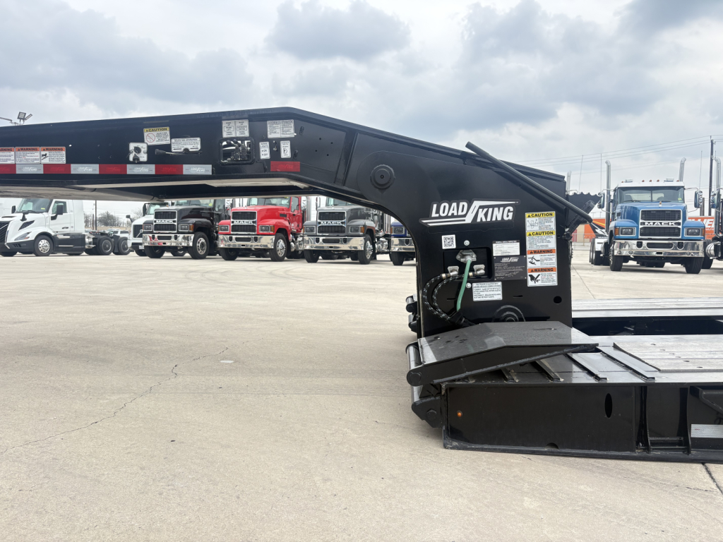Load King 503/605-8'-6"-20" lowboy trailer arm and platform showing hydraulic winch area and warning labels, with a row of trucks in the background.