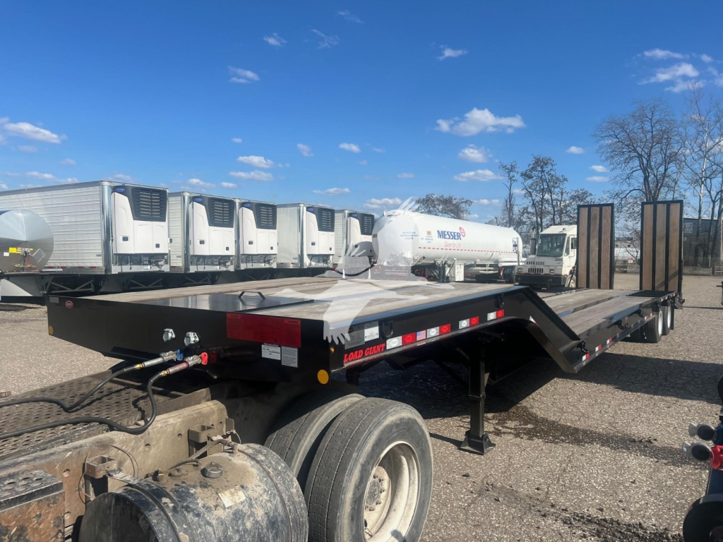 Trailer deck showing loading ramps and dual rear axles with flatbed surface, black frame, red and white reflective tape, and various equipment in background such as fuel tanks and trailers.