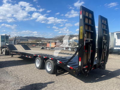 Trailer in a parking lot showing a black drop deck trailer with rear loading ramps raised, dual rear axles, and a long low-profile flatbed.