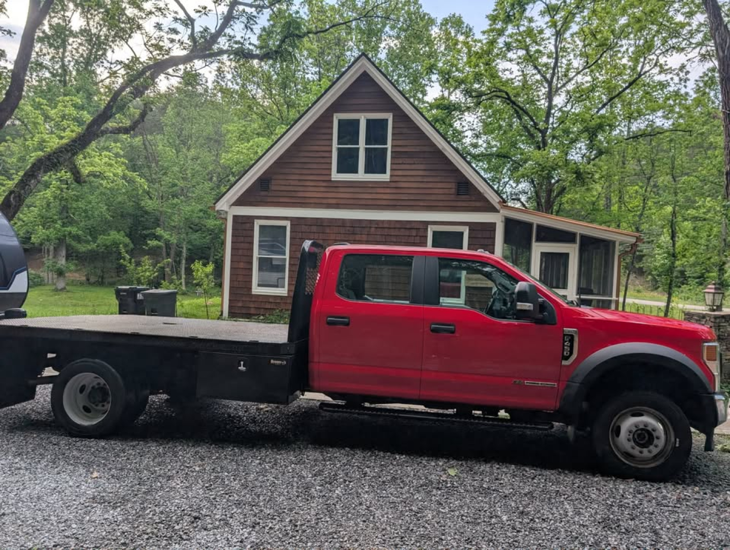 Ford F-450 Super Duty XL flatbed truck side profile in red with a gooseneck flatbed, 4-door crew cab, on a gravel driveway in front of a wooden house.