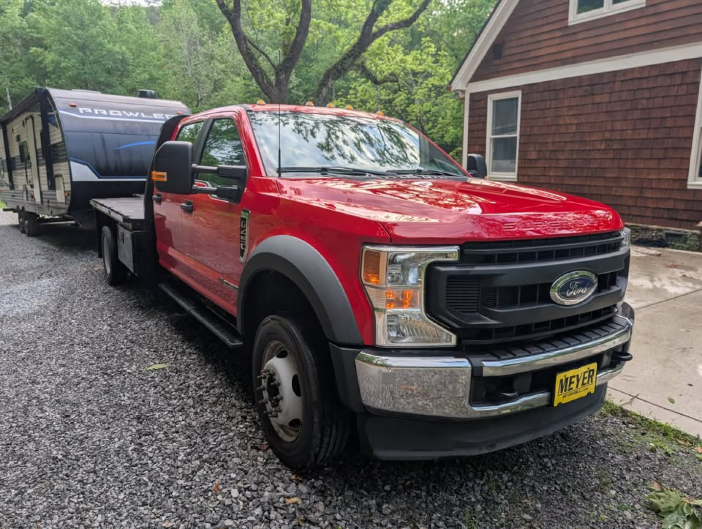 Ford F-450 Super Duty XL truck with flatbed and a trailer in the background; front grille shows Ford emblem, parking on a gravel surface, yellow dealer plate.