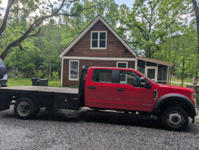 Ford F-450 Super Duty XL flatbed truck side profile in red with a gooseneck flatbed, 4-door crew cab, on a gravel driveway in front of a wooden house.