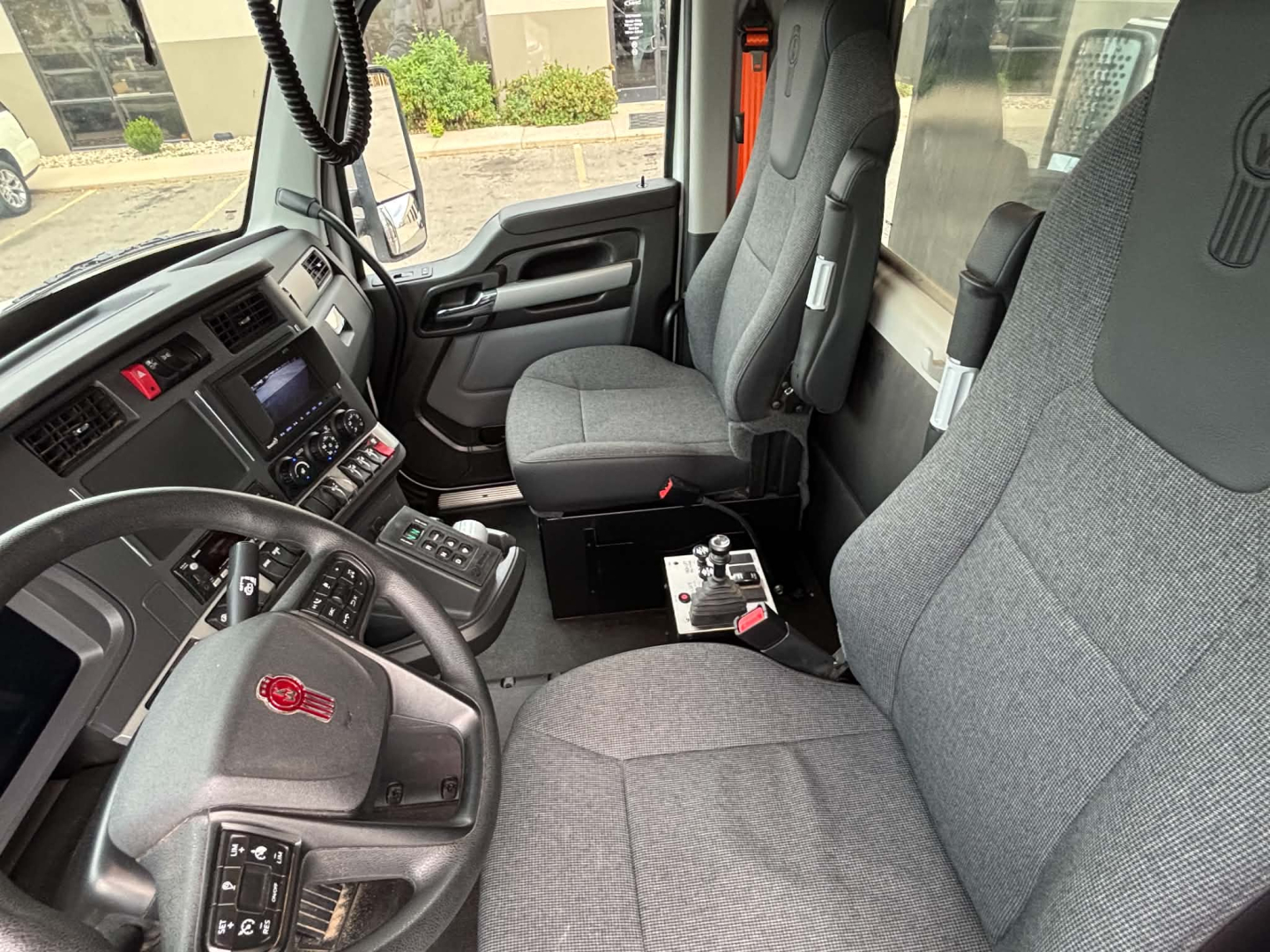 Interior of Kenworth T880 cab showing steering wheel, dashboard, and grey cloth seats. Center console with controls, passenger seat, and gear area.