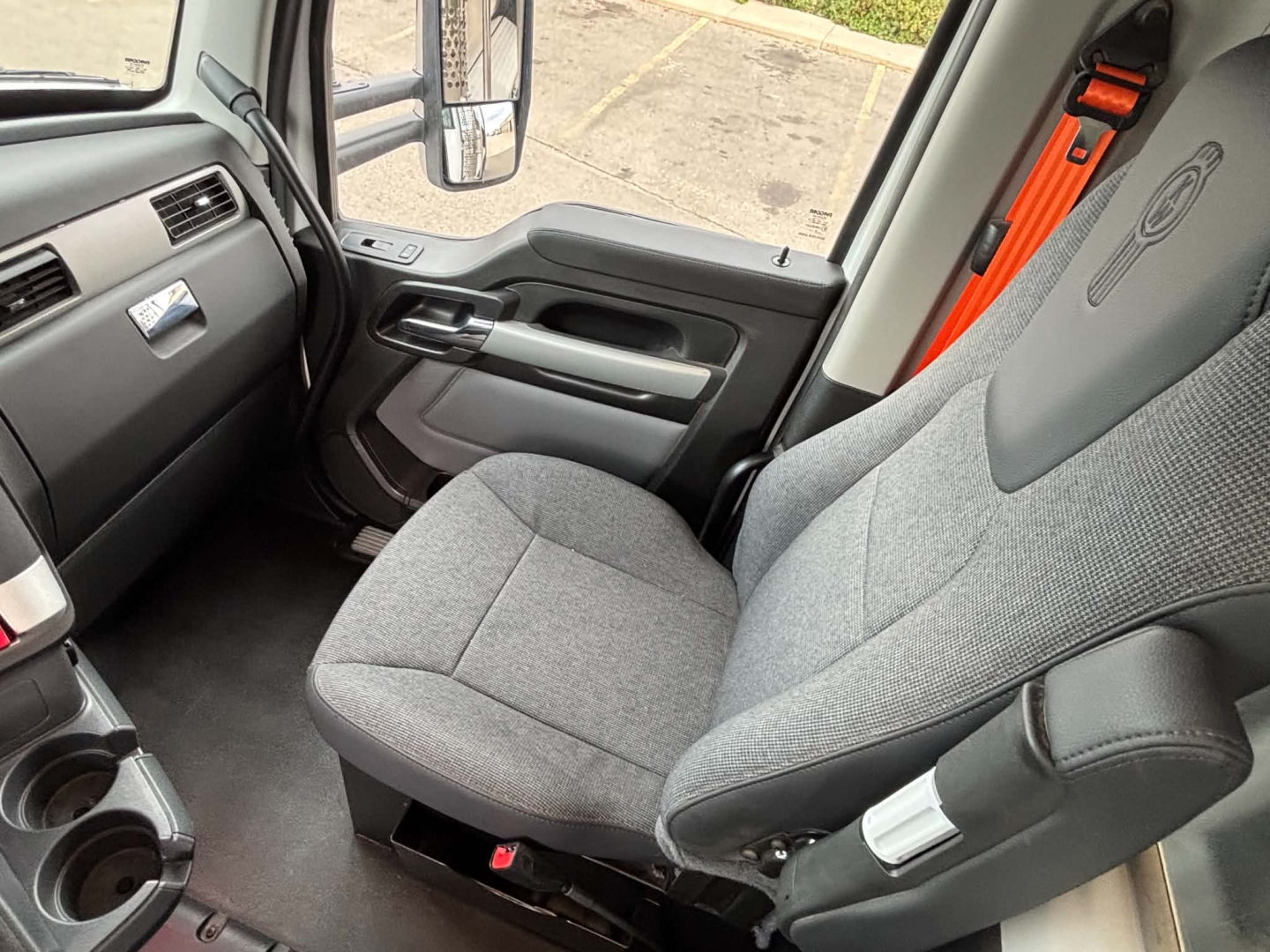 Cab interior of a Kenworth T880 truck showing gray fabric driver seat, dashboard, steering wheel, door panel, and orange seat belt in view.