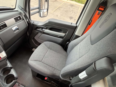 Cab interior of a Kenworth T880 truck showing gray fabric driver seat, dashboard, steering wheel, door panel, and orange seat belt in view.