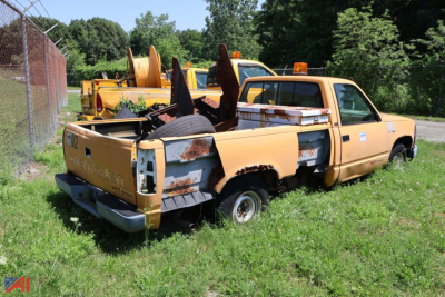 Chevrolet C/K 1500 pickup truck in salvage yard with rust and missing bed, yellow paint, rear view showing tailgate and damaged bed sides.
