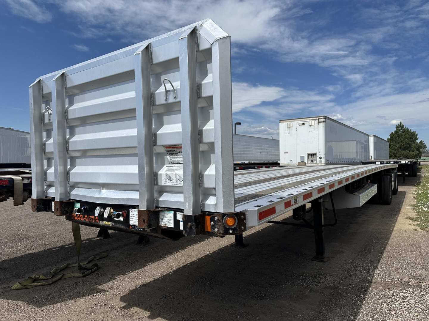 Fontaine Infinity flatbed trailer with tall rear ramp gate, aluminum sides, and tandem axles seen in a gravel lot under blue sky.