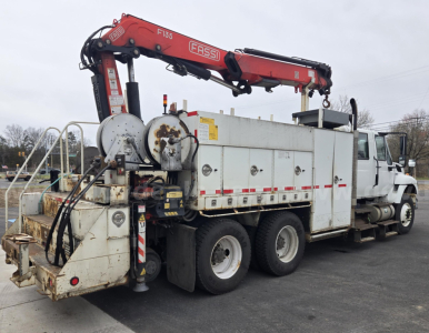 Truck-mounted crane on a white service body with multiple compartments; red Fassi crane labeled F135 on top; various hoses and gauges at rear; worn metal platforms and tires visible.