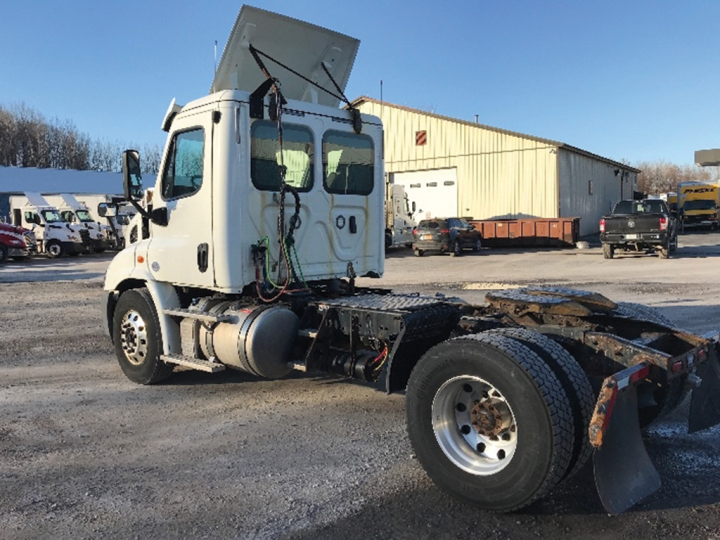 White Freightliner Cascadia daycab tractor with rear chassis and exposed fifth wheel, cab doors, and fuel tanks visible; background shows a truck yard.