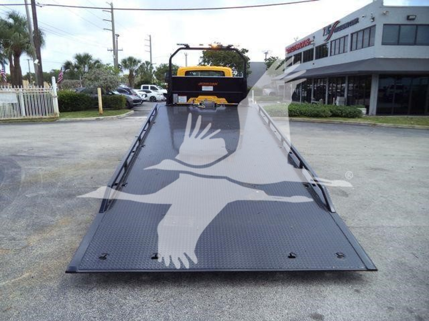 Truck with a flatbed showing a large white bird graphic inservice. The bed is metallic with tie-down points; rear of a rollback truck visible in distance.
