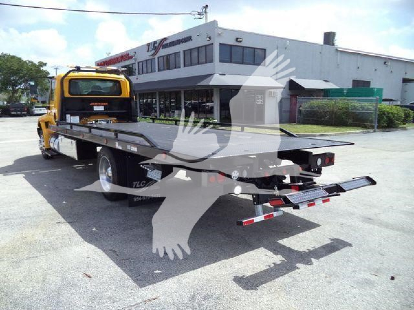 Flatbed tow truck on a dealership lot with a yellow cab and long black bed; building in background.