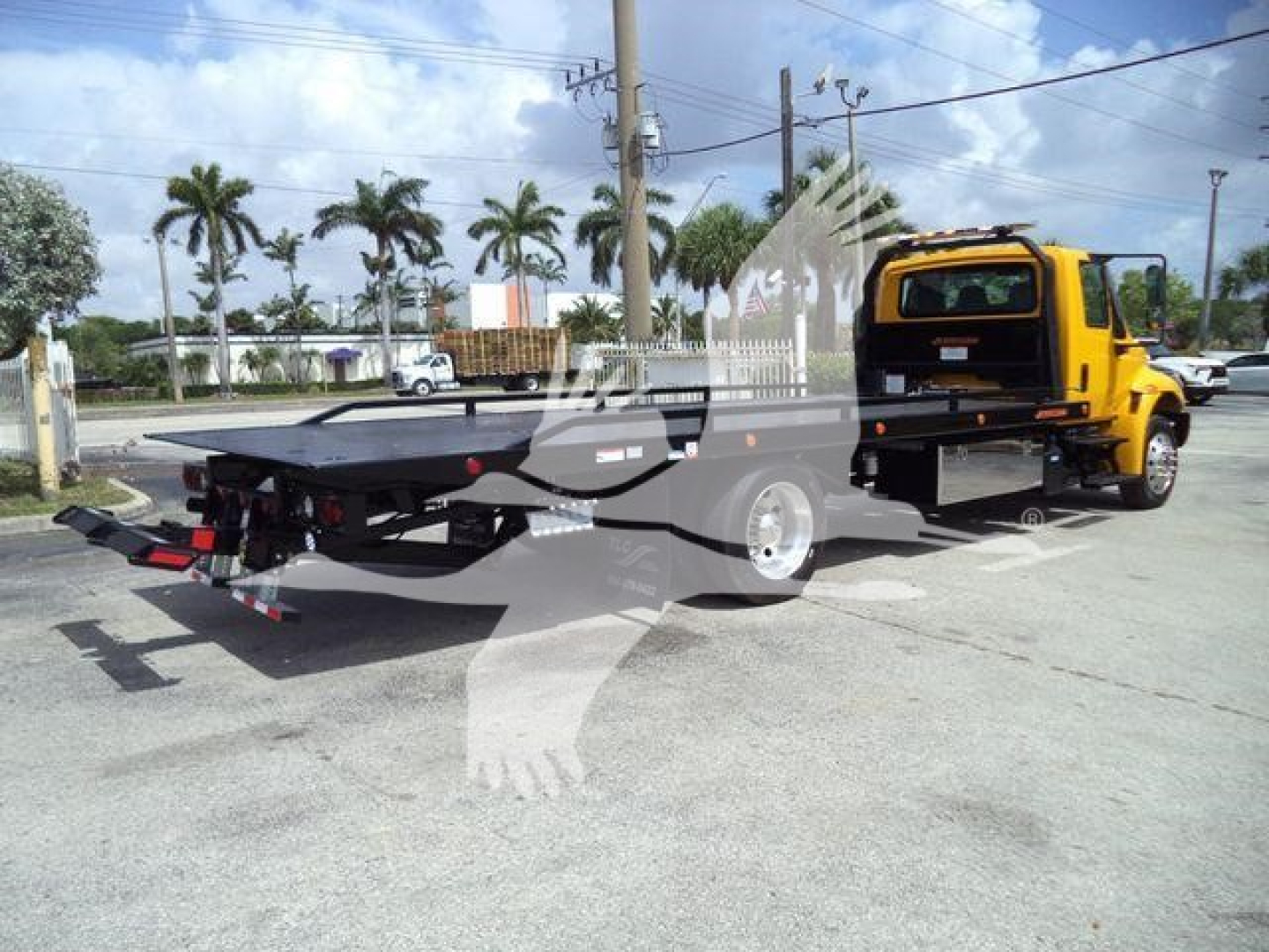 Flatbed tow truck with yellow cab showing rear flatbed and hooks, parked in lot.