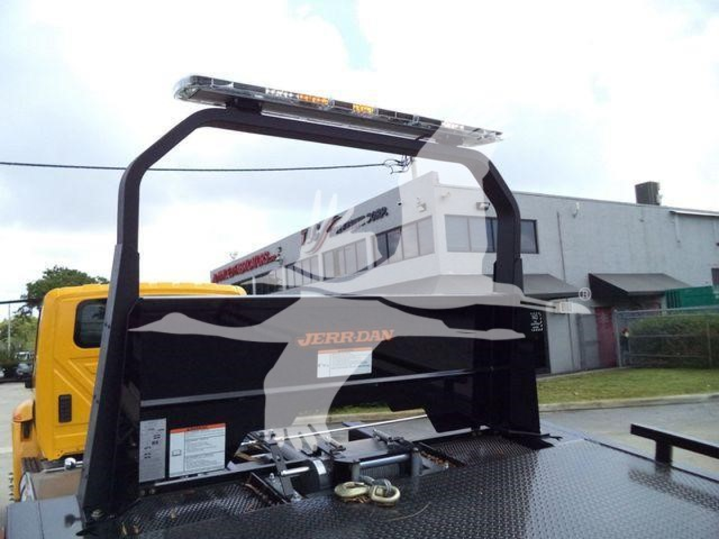 Truck rollback bed with black roll-over protection arch and warning lights; white cab in background, JERR-DAN branding on bed, metal diamond plate surface.