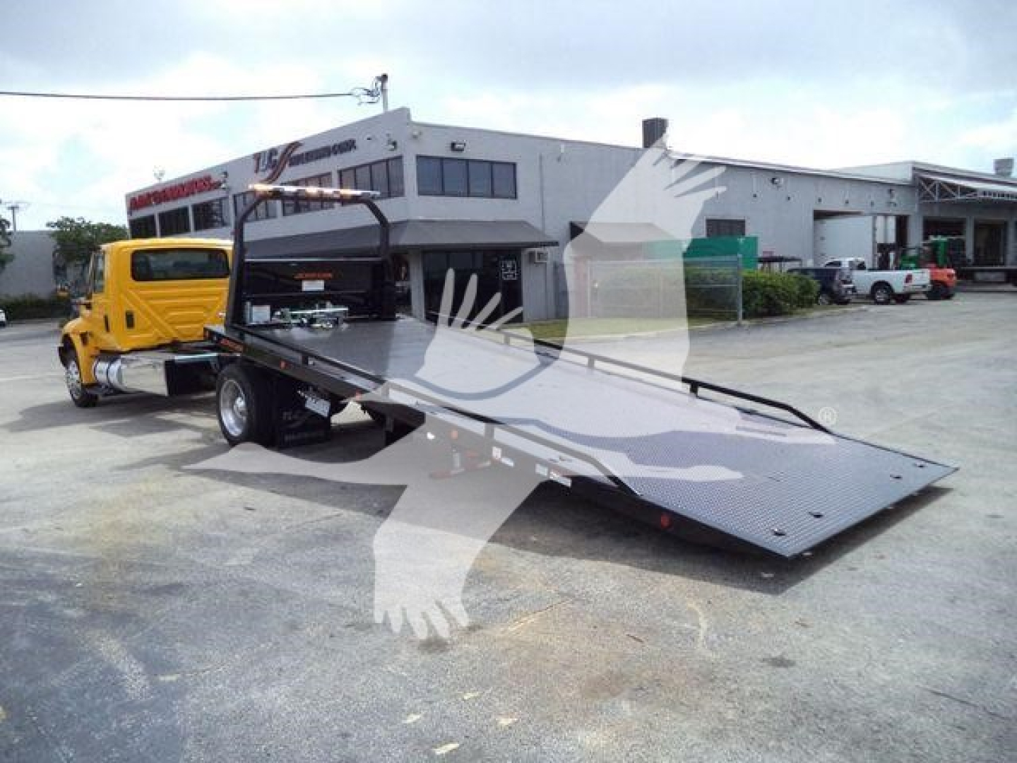 2019 International 4300 rollback truck with flatbed extends forward; yellow tow truck in background; dealership lot with building in distance.