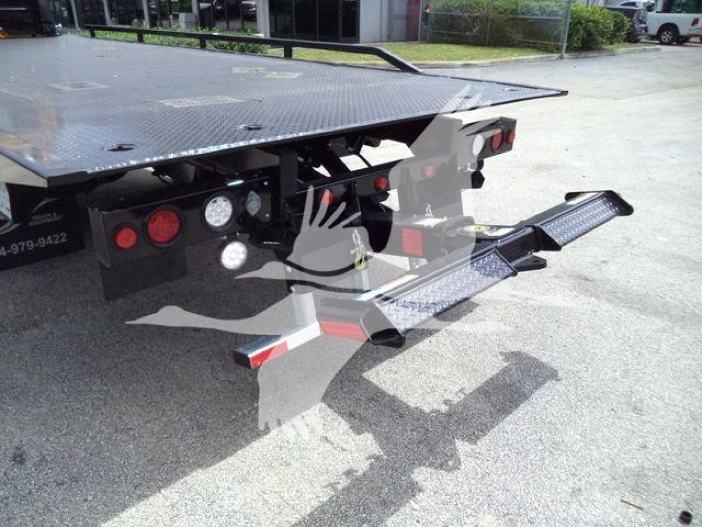 Rear of a flatbed tow truck showing a hydraulic stowable bed and wheel lift with red and white reflectors. Visible tags and lights on rear frame.