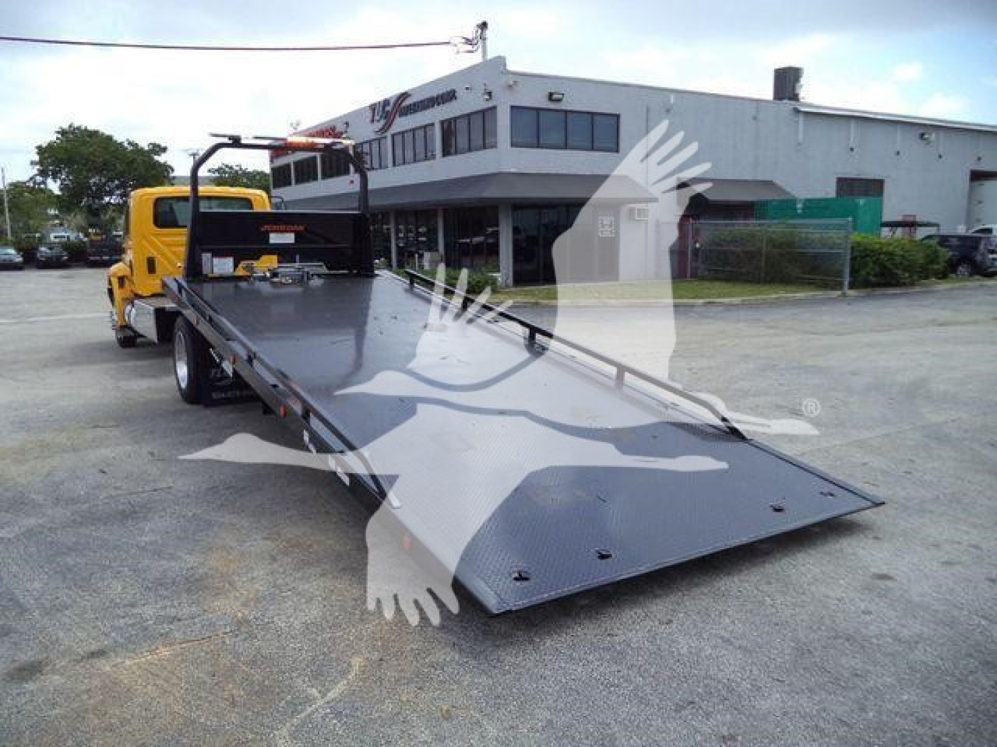 Flatbed rollback tow truck with a long steel bed, yellow cab, and black ramp lying in a dealership lot.