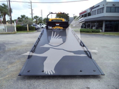 Truck with a flatbed showing a large white bird graphic inservice. The bed is metallic with tie-down points; rear of a rollback truck visible in distance.