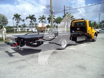 Flatbed tow truck with yellow cab showing rear flatbed and hooks, parked in lot.