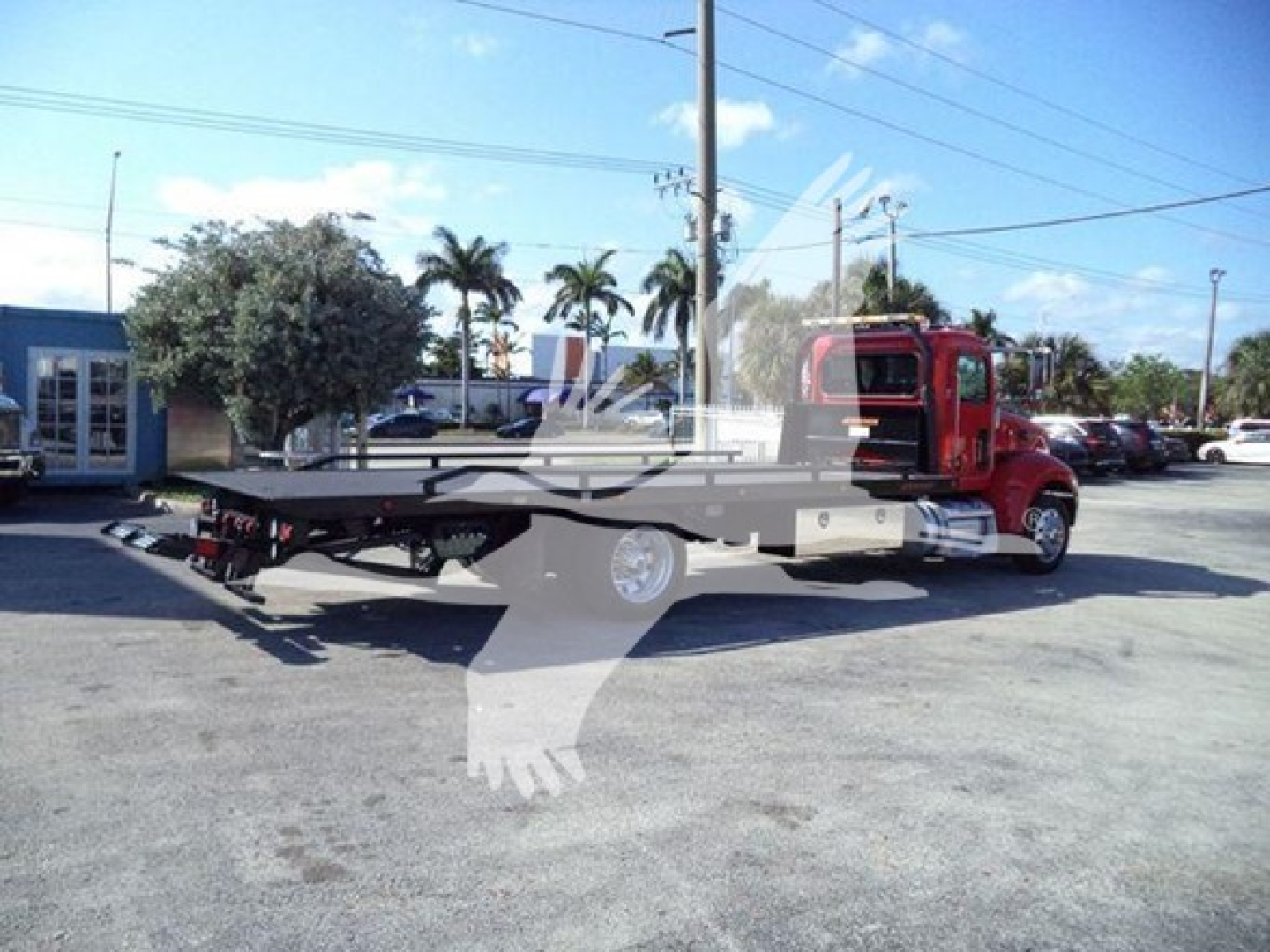 Peterbilt 337 rollback tow truck in side view showing red cab and flatbed with equipment; background includes palm trees and dealership lot.