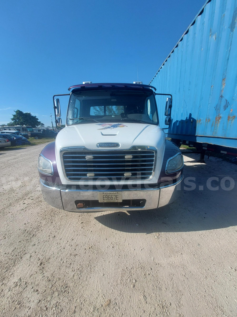 Front view of a white Freightliner M2 truck with a large grille, bumper, and license plate. Blue trailer on the right, clear sky.