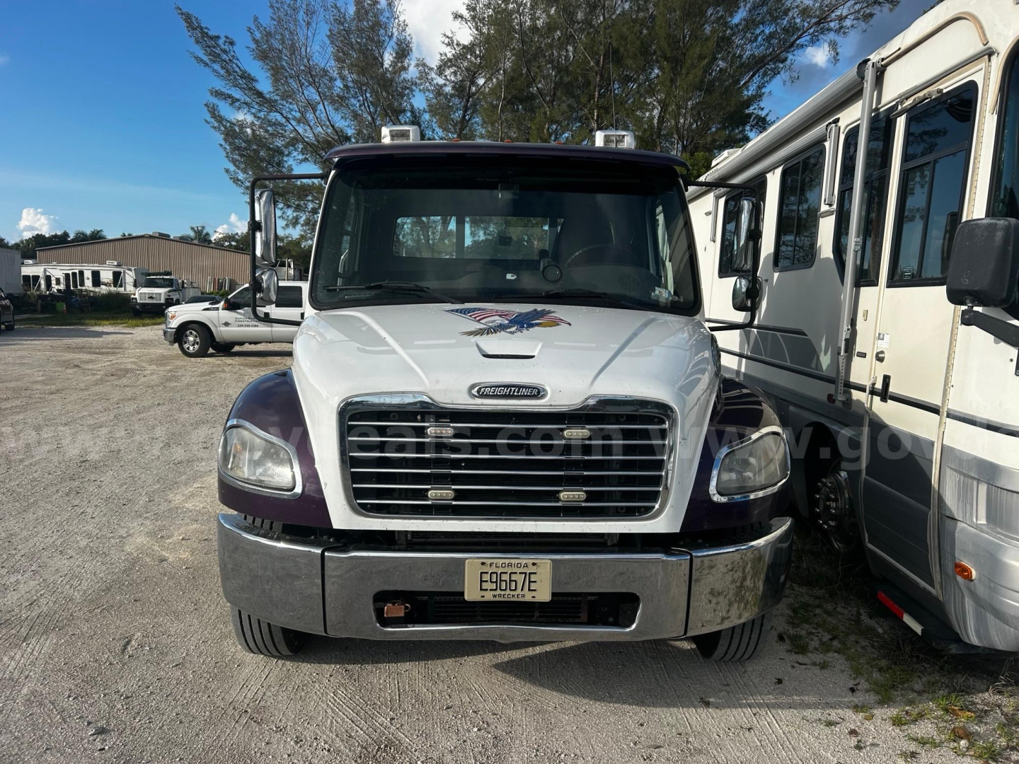 Front view of a 2019 Freightliner M2 tow truck with a white cab, chrome grille, and yellow license plate E9676E. Emblem on hood, windshield wipers, and passenger side mirrors visible.