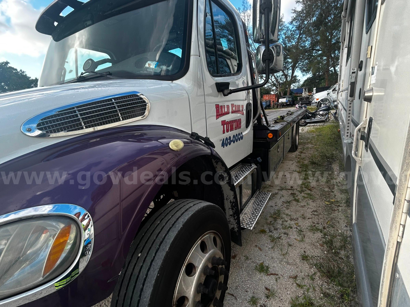 Freightliner M2 tow truck side view showing cab with purple fender and white cab, dual rear mirrors, steps, and side logo.