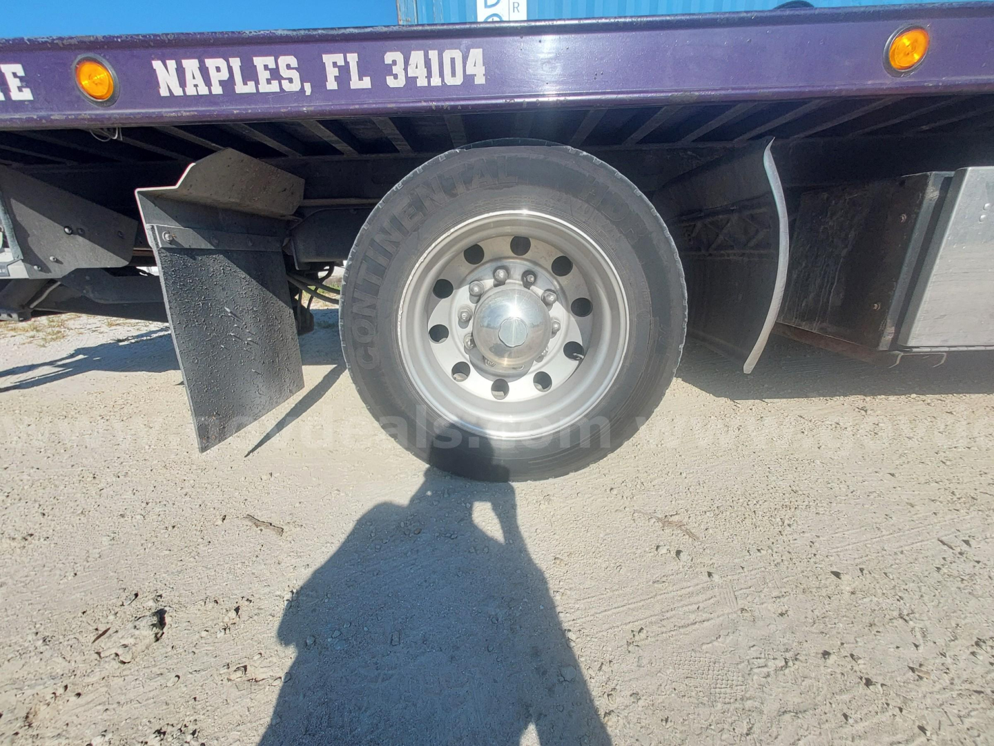 Truck wheel and rear fender area of a Freightliner M2 with Naples, FL 34104 text on the bed, showing a single rear wheel, mud flaps, and a placeholder shadow.