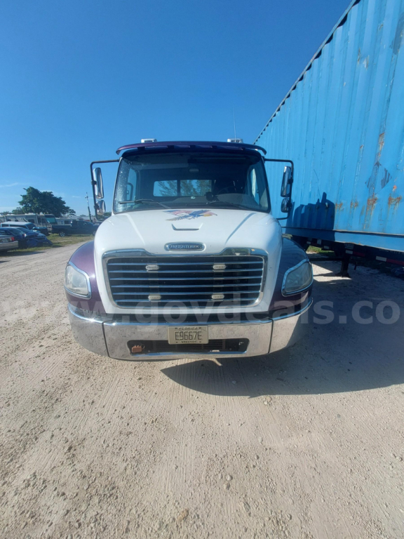 Front view of a white Freightliner M2 truck with a large grille, bumper, and license plate. Blue trailer on the right, clear sky.