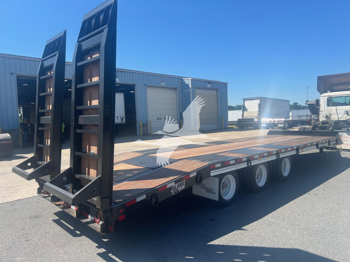 Trailer in service bay showing low-bed flatbed with dual ramps and multiple axles; black uprights at rear, wooden deck, multiple wheels; blue sky overhead.